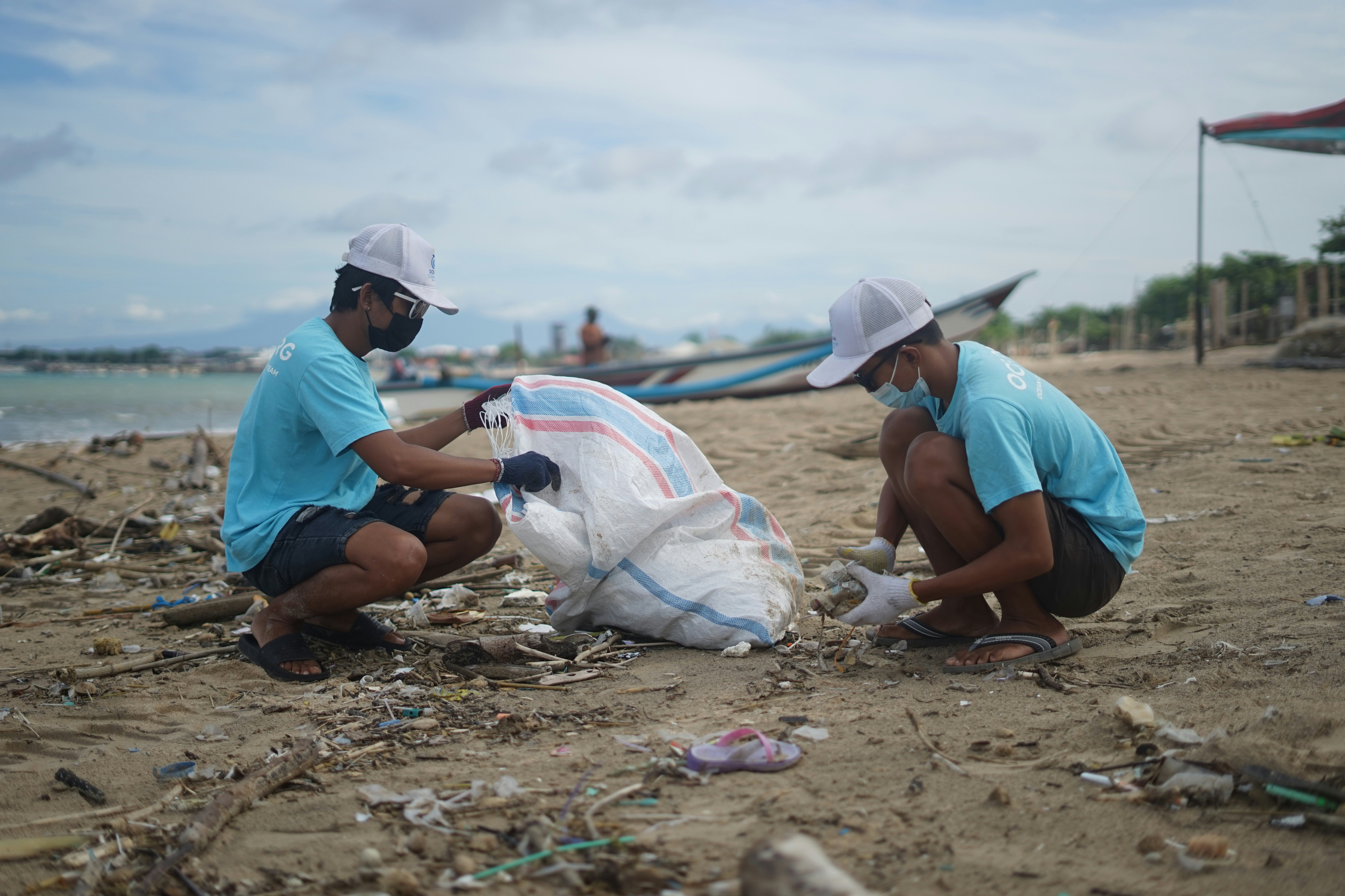Two Indonesian males clean the beach on behalf of ocean cleanup group. OCG is a free ocean search engine that cleans the ocean. by Gone Greenish man in blue shirt sitting on sand during daytime
