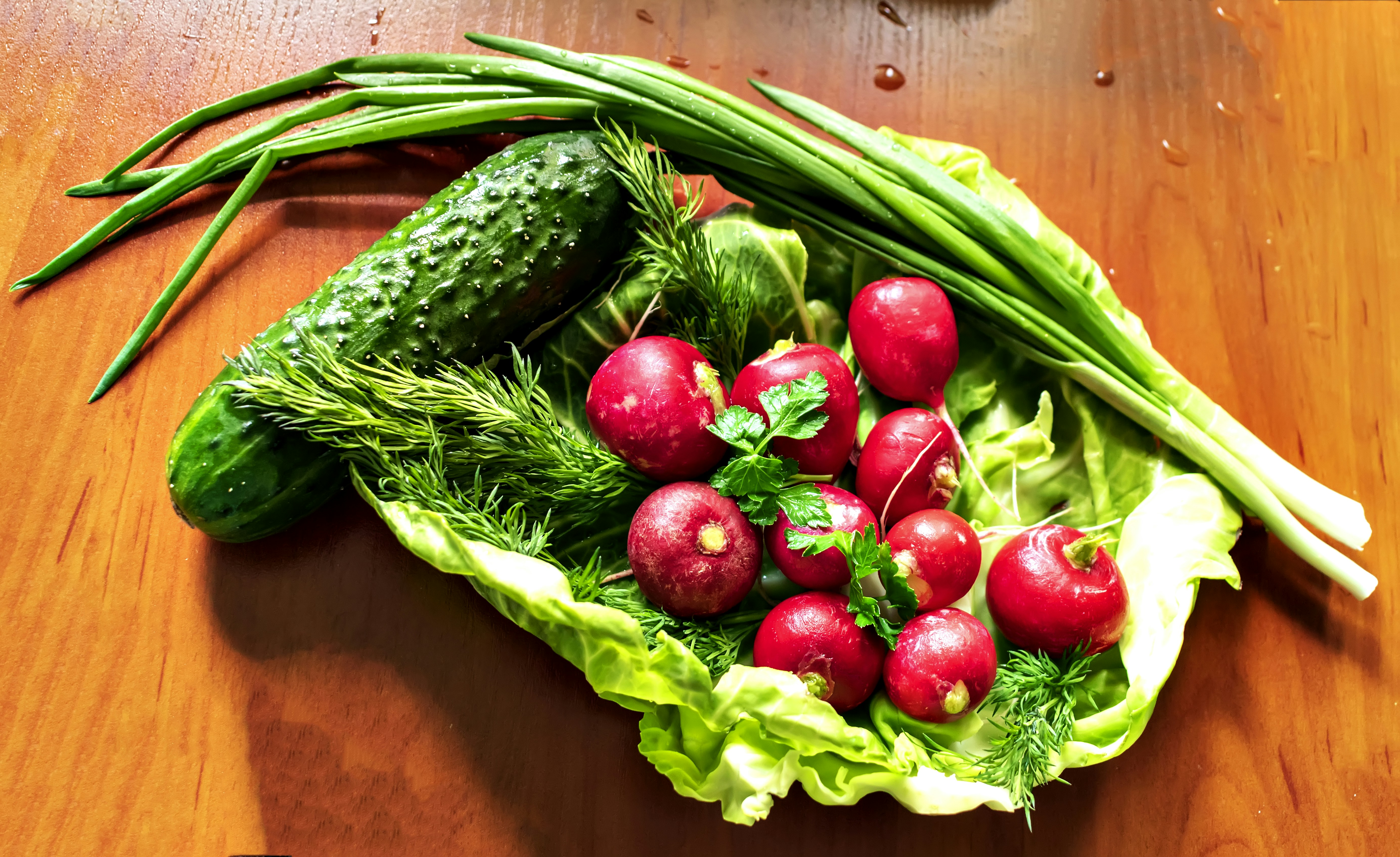 There are a few radishes laying on a cabbage leaf on the table. Add cucumber, green onion, parsley and dill. by Gone Greenish a wooden table topped with lettuce, radishes and cucumbers