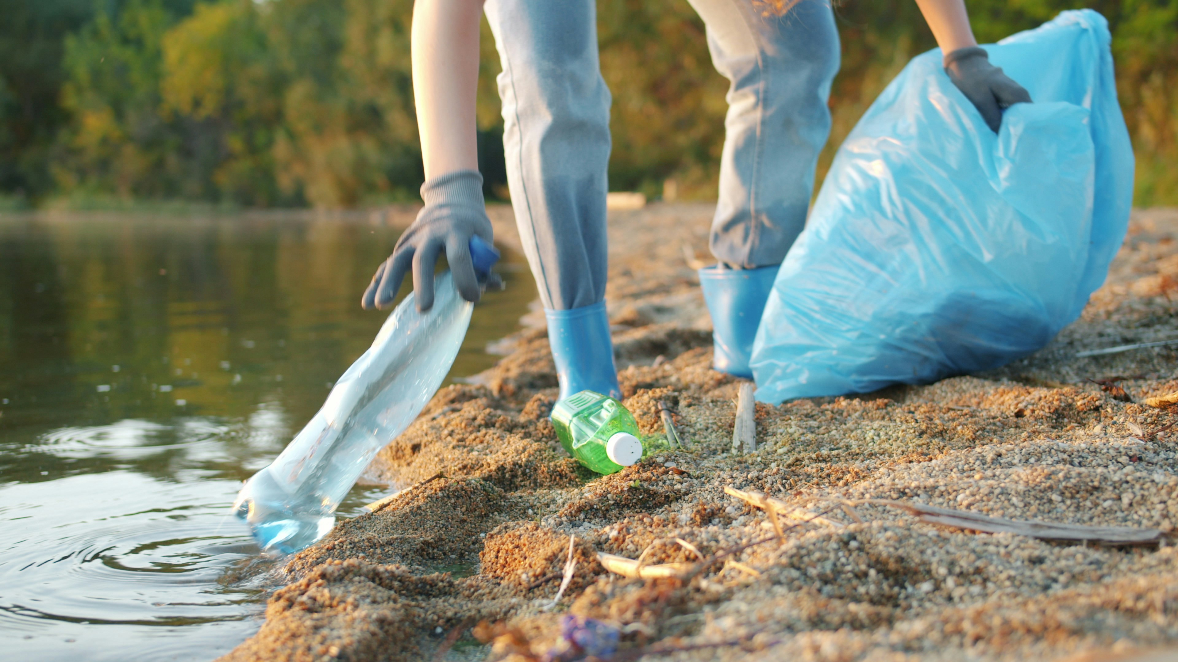 Close-up of female legs and hands collecting garbage on the lake shore in summer, female volunteer wearing gloves and rubber boots is cleaning nature. by Gone Greenish Person picking up plastic bottle near water