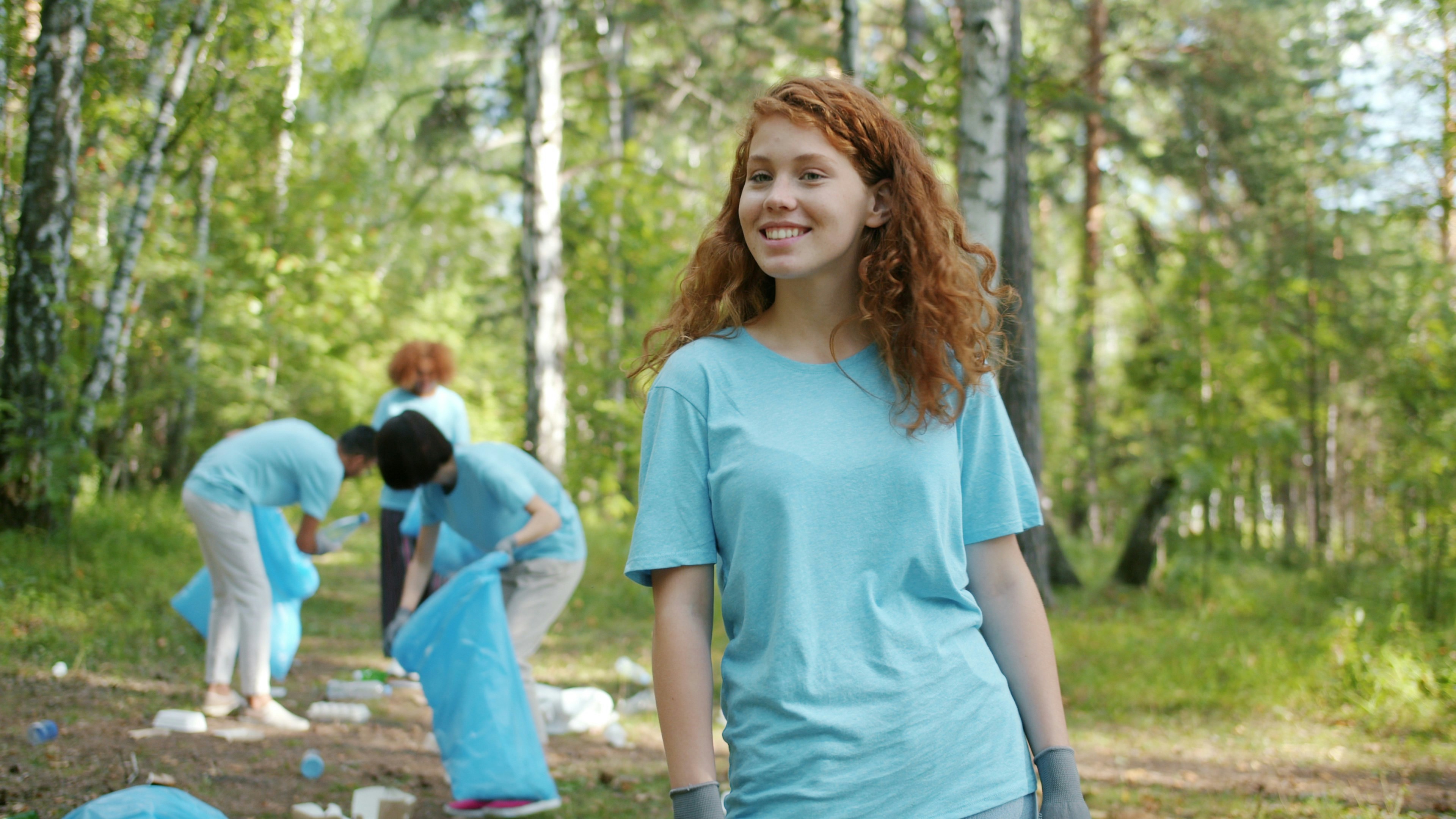 Portrait of young female eco activist smiling in forest while team of eco-friendly people is collecting trash in bib bags. Nature, students and environment concept. by Gone Greenish Young woman smiles while cleaning forest with volunteers