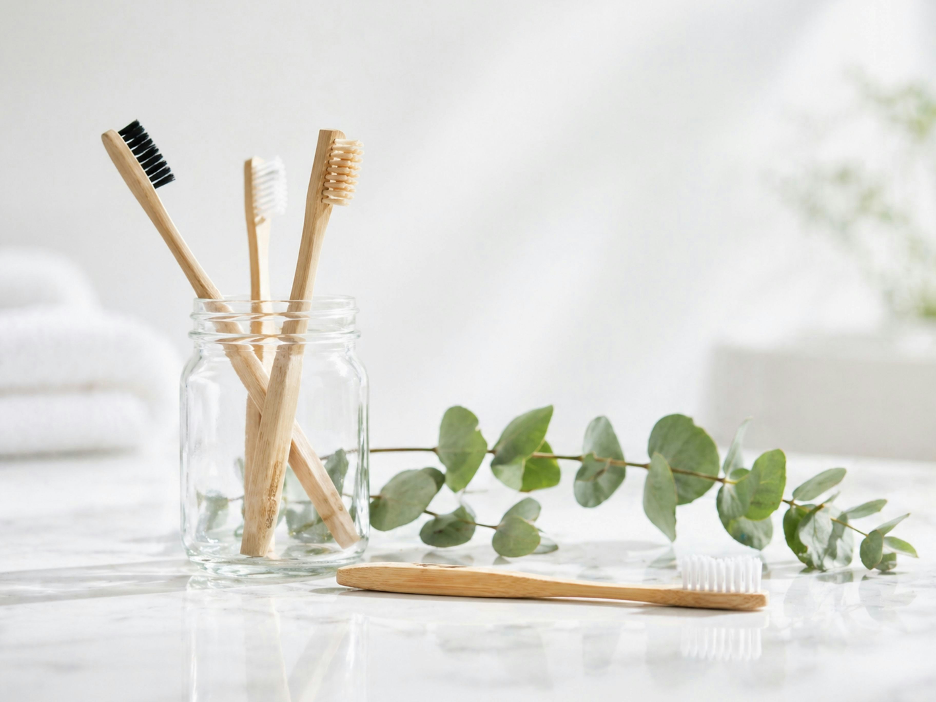 A clean, minimalist scene featuring bamboo toothbrushes in a glass jar, highlighting sustainable dental hygiene and eco-friendly oral care in a bright, calming setting. by Gone Greenish Bamboo toothbrushes in a glass jar with eucalyptus leaves.